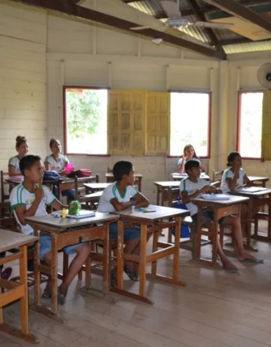 Crianças indígenas. Elas estão uniformizadas, em uma sala de aula. A estrutura do local é simples e as mesas são de madeira.